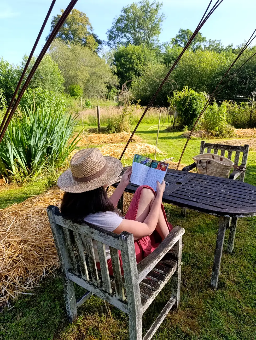 Ysabel en chapeau de paille, lisant à la table de son jardin en permaculture